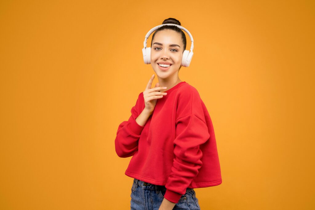 Smiling young woman in red sweatshirt listening to music with wireless headphones against a vibrant background.